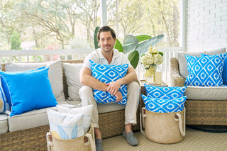 A man relaxes on a wicker sofa with the Blue Ikat Pillow on a sunny porch, surrounded by vibrant blue-patterned pillows, woven baskets, and white flowers, enjoying the modern charm and view of trees outside.