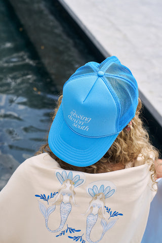 A person in a blue Sewing Down South Trucker hat from The Siren Collection is seen from the back. Their curly hair complements a cream-colored shirt featuring two stylized mermaids. The hats adjustable snapback and cotton blend ensure comfort by the poolside.