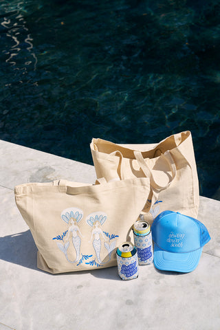 Two beige tote bags with blue, white, and floral designs sit by a pool. Nearby are three canned drinks in SDS Calico Mini Floral Koozies and a blue cap with white text. The sunny scene highlights the pools deep blue waters.