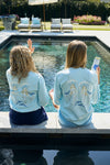 Two people sit on the edge of a pool in matching SDS Sirens Long Sleeve Tees from The Siren Collection. One waves while the other holds a smartphone. Their retro-style light blue shirts with mermaid designs complement the lounge chairs and greenery in the background.