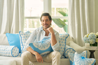 A smiling man in a light shirt sits on a white sofa, holding the Calico Scallop Lumbar Pillow with blue floral patterns. Matching blue and white cushions, large windows, white curtains, and a potted plant create a sophisticated backdrop.