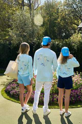 Three individuals in matching light blue Siren Collection: SDS Sirens Long Sleeve Tees and blue caps face a garden, admiring the flower beds with sunlight filtering through trees, creating a serene, retro-style scene.