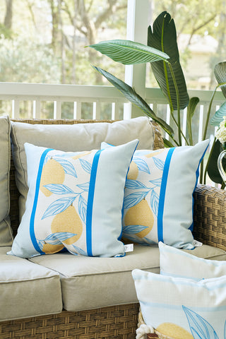 Two Lemons in a Line Pillows with a yellow lemon and blue leaf design sit on a beige wicker outdoor sofa. A green plant and white railing complete this cozy, sunlit patio, ideal for citrus-themed decor.
