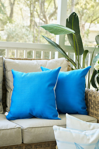 A Lemons in a Line Pillow sits on a beige outdoor wicker sofa, surrounded by green leafy plants and soft window light, creating a cozy and inviting patio atmosphere.