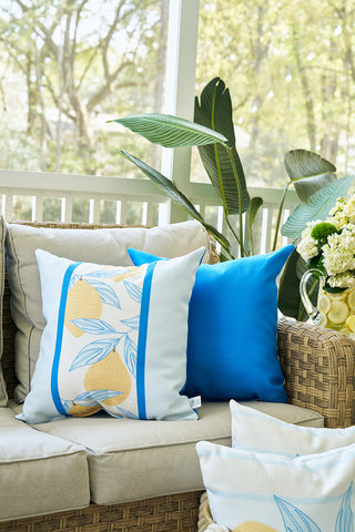 A cozy wicker sofa with beige cushions features the Lemons in a Line Pillow and blue decor. Large green plants and a vase of flowers complete the inviting, sunlit porch.
