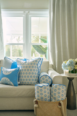 A cozy living room features a cream sofa with blue and white patterned pillows, a woven basket holding the Calico Mini Floral Bolster Pillow, and a side table with vibrant blue flowers by a bright window.