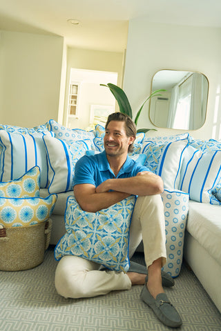 A smiling man in a blue shirt sits barefoot on a white couch with Euro Blue Tiles with Spring Blue Stripe Pillow, holding it in his arms. Neutral walls and a mirror behind him complete the room’s European charm.
