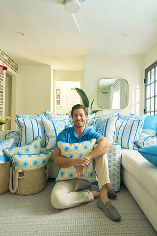 A smiling man in a blue shirt and beige pants sits on the floor of a bright living room, surrounded by blue and white patterned pillows and the Yellow Calico Scallops Pillow, creating a relaxing coastal vibe. More pillows decorate the white couch behind him.