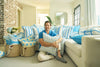 A smiling man sits on the floor of a bright living room, surrounded by blue and white pillows, including a Calico Mini Floral Oversized Pillow with bright blue piping. Baskets filled with pillows and flowers add to the inviting, cozy space.