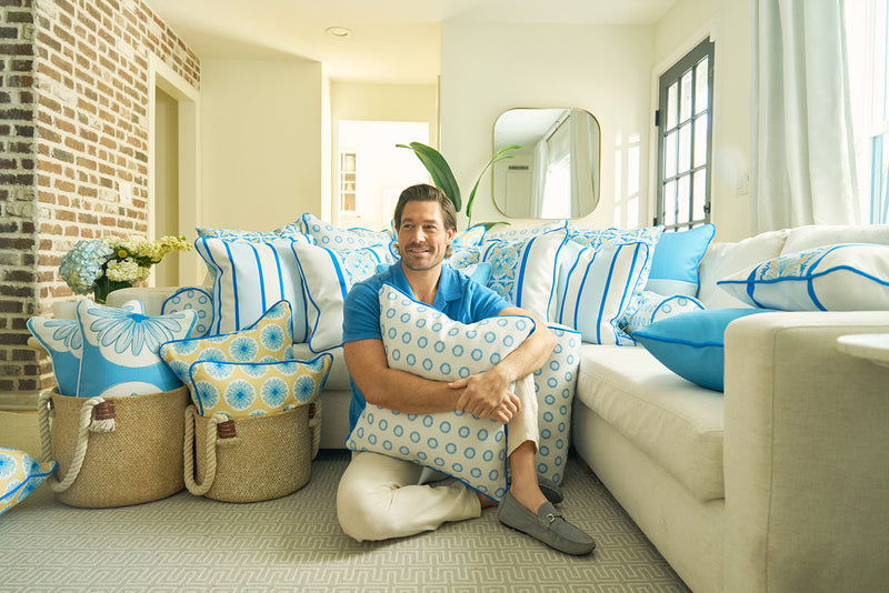 A smiling man sits on the floor of a bright living room, surrounded by blue and white pillows, including a Calico Mini Floral Oversized Pillow with bright blue piping. Baskets filled with pillows and flowers add to the inviting, cozy space.