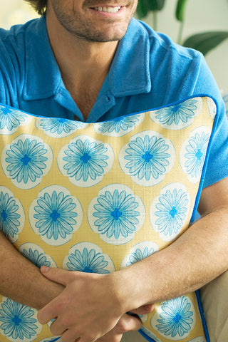 A person in a blue shirt smiles while hugging the Yellow Calico Scallops Pillow, featuring blue and white floral patterns. The softly lit background with greenery enhances the cozy coastal feel.