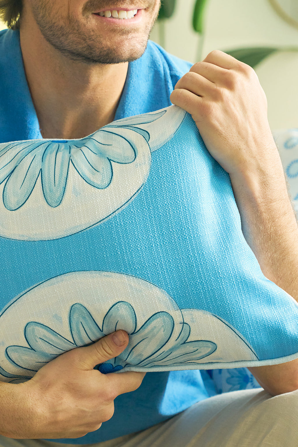 A man in a blue shirt hugs the Calico Scallop Lumbar Pillow, featuring white and blue floral designs. Only his smiling mouth and hands are visible, evoking a sophisticated, coastal summer vibe.