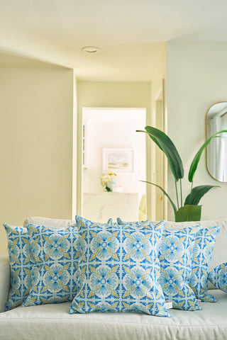The Euro Blue Tiles Pillow, with blue and yellow floral accents, brightens a modern living room’s light sofa alongside other stylish throw pillows, complemented by a background of greenery and wall art.