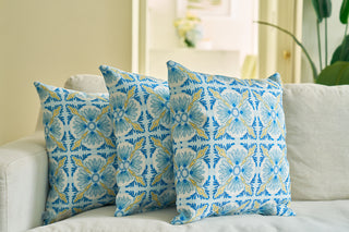 The Euro Blue Tiles Pillow, featuring a striking floral design, is arranged with two other decorative pillows on a light-colored sofa in a bright, cozy living room with a blurred plant and vase of flowers in the background.