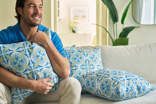 A man sits on a light-colored sofa, smiling and hugging the Euro Blue Tiles Pillow. More accent pillows are beside him, with green plants adding fresh decor to the bright, modern living room.