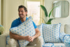 A man sits on a white couch, smiling and hugging a Calico Mini Floral Oversized Pillow with bright blue piping, surrounded by matching cushions in a bright, modern living room.