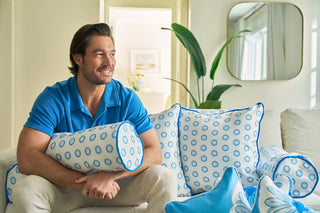 A smiling man in a blue shirt sits on a light sofa, holding the Calico Mini Floral Bolster Pillow. Coordinating pillows with vivid blue flowers surround him, while a large green plant and mirror decorate the background.