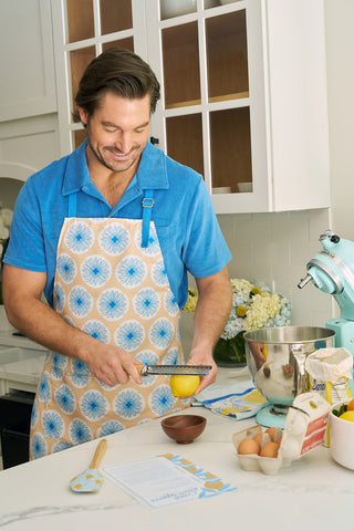A man in a blue shirt and a Calico Scallops on Yellow Apron with an adjustable neck strap zests a lemon over a bowl in the kitchen. Eggs, a recipe card, and a Sewing Down South stand mixer are on the counter as he smiles.