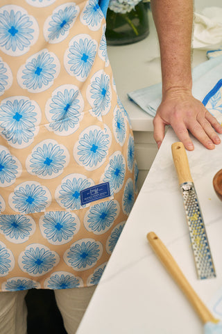 Wearing a Calico Scallops on Yellow Apron with an adjustable neck strap, a person stands by the kitchen counter resting a hand beside a grater and wooden-handled knife, with a blue and beige cloth nearby.