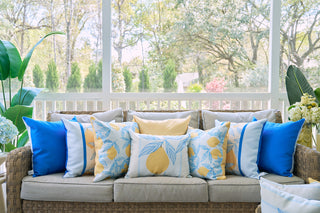 A wicker sofa with tan cushions is styled with blue, white, and yellow accent pillows, including the Lemons in a Line Pillow for a citrus touch. Green plants and outdoor trees are visible through the screened porch behind the sofa.