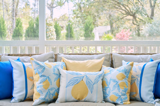 A cozy outdoor sofa features the Lemons in a Line Lumbar Pillow and blue and white cushions, adding citrus-inspired decor to a porch with a white railing overlooking a lush green garden.
