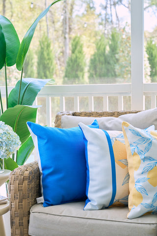 A wicker sofa with three decorative pillows—including the Lemons in a Line Pillow—on a porch, surrounded by green plants and soft natural light.