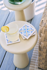 A glass of lemonade with a lemon slice rests on a white table beside the Euro Blue Tile Coaster Set, next to a woven chair and beige rug on a wooden deck.