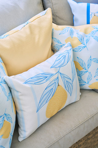 A group of decorative pillows on a beige sofa, including the Lemons in a Line Lumbar Pillow, features yellow and white fabrics with blue-outlined leaves and yellow citrus fruit patterns for a fresh, citrus-inspired decor.