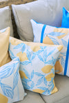 Four pillows brighten this cozy corner on a light beige sofa. The Lots of Lemons Pillow and its blue leaf companion have vibrant designs, while the other two are solid yellow and white with blue accents. A wicker backrest peeks behind them.