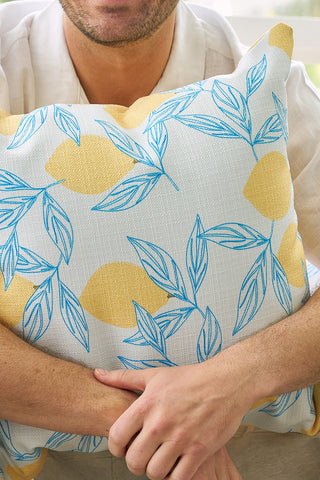 A person in a light-colored shirt hugs the Lots of Lemons Pillow, featuring a white background with blue leaf details and yellow abstract shapes—an ideal accent to brighten any cozy nook. Only their lower face and arms are visible.