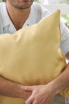 A person with light facial hair and a light shirt hugs a large Lots of Lemons Pillow to their chest in a cozy, softly lit corner, creating a relaxed and inviting atmosphere.
