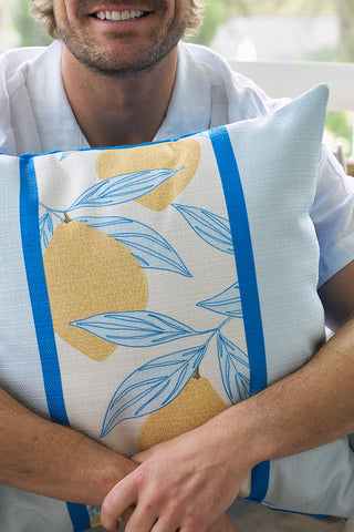A smiling man in a light shirt holds the Lemons in a Line Pillow, featuring yellow lemons, blue leaves, and blue stripes. The softly lit background is out of focus.