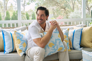 A man with brown hair and a light beard sits on a sofa, smiling as he holds a vibrant Lots of Lemons Pillow, surrounded by other patterned pillows in a cozy, sunlit patio corner with trees visible outside.