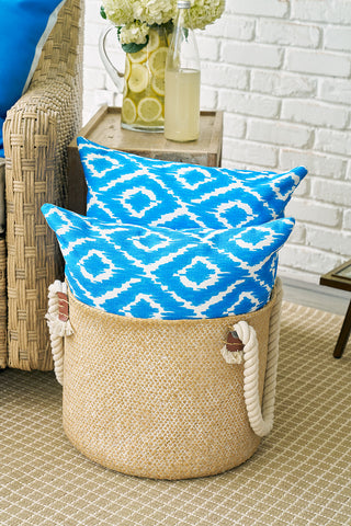 A woven basket with rope handles holds two Blue Ikat Pillows, creating a modern, timeless look. The basket sits on a beige rug next to a wicker chair and a wooden side table topped with a lemon drink and white flowers.