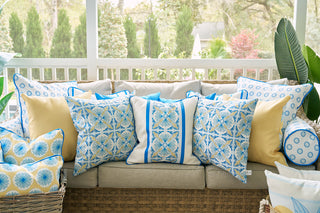 A wicker sofa on the porch is adorned with blue, yellow, and Calico Mini Floral throw pillows—including a cozy Calico Mini Floral Bolster Pillow—set amid leafy plants and a screened outdoor view.