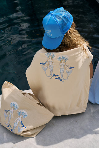 A person with curly hair wears a blue cap and a cream The Siren Collection: SDS Sirens Hoodie with mermaid art, paired with a matching cream tote by the pool, bringing coastal fun to the scene.