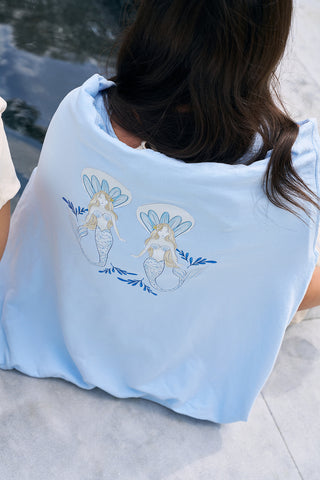 A person with long hair sits by a pool wearing The Siren Collection: SDS Sirens Hoodie in light blue, featuring two crowned mermaids surrounded by small blue leaves on the back. The relaxed fit is crafted from soft ring-spun cotton, adding charm.