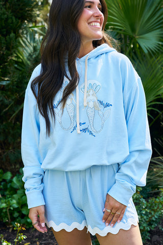 A woman smiles outdoors in a light blue SDS Sirens Hoodie from The Siren Collection, featuring floral and mermaid designs, paired with matching wavy-hem shorts. Green foliage and palm leaves create a vibrant, coastal vibe.