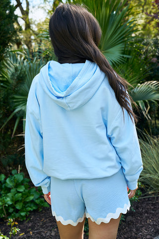 A person with long hair stands outdoors, seen from behind, wearing The Siren Collection: SDS Sirens Hoodie in light blue and matching relaxed fit shorts featuring a scalloped hem. Lush greenery and plants form the background.