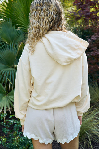 A person with curly hair, wearing The Siren Collection: SDS Sirens Hoodie in a relaxed fit light beige style, stands outdoors among lush greenery and a tree with burgundy leaves.