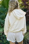 A person with curly hair, wearing The Siren Collection: SDS Sirens Hoodie in a relaxed fit light beige style, stands outdoors among lush greenery and a tree with burgundy leaves.