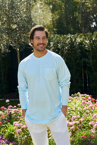 Dressed in The Siren Collections heavyweight, unisex relaxed fit long sleeve tee and white pants, a person smiles amidst a garden filled with pink flowers. Sunlight filters through the trees in the background, crafting a serene atmosphere.