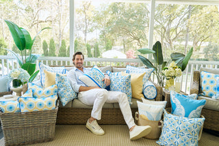 A man relaxes on a sofa in a bright sunroom, surrounded by Line of Euro Blue Tiles Pillows. Green plants and a basket of white flowers bring European elegance, creating a cozy and inviting atmosphere.