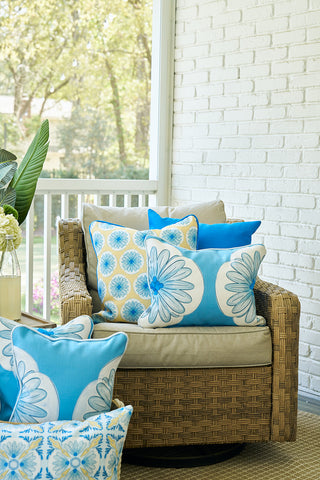 A wicker armchair with beige cushions and the Calico Scallop Lumbar Pillow adds sophistication, while blue and white floral throws enhance coastal charm on a porch with a white brick wall and lush potted plants in the background.