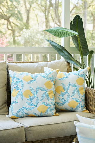 Two beige outdoor sofa cushions and the Lots of Lemons Pillow with blue leaf prints rest on a wicker couch. A large green plant and white porch railing in the background create a cozy corner.