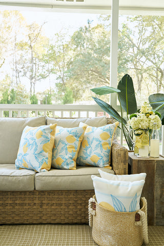 A cozy nook features a wicker patio sofa with the Lots of Lemons Pillow, leaf-patterned pillows, and a rug. Nearby, a basket holds extra pillows and a side table displays flowers, lemonade, and a leafy plant. Trees can be seen outside.