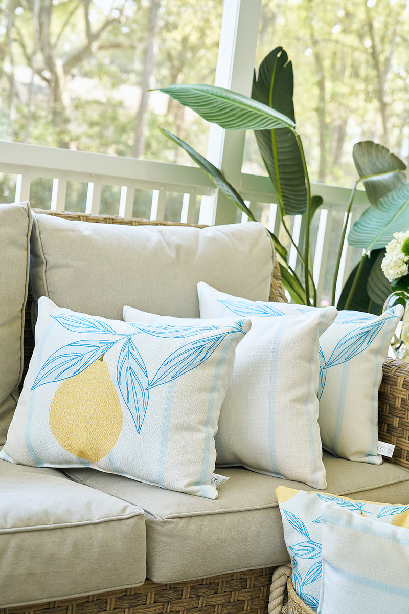 A wicker sofa with light beige cushions features white pillows with blue leaves and a Lemons in a Line Lumbar Pillow. A large leafy plant and white railing in the sunlit background enhance the citrus-inspired decor.