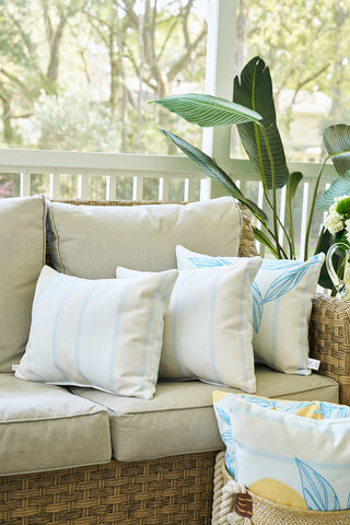 A beige wicker outdoor sofa with light tan cushions is accented by white, light blue, and the Lemons in a Line Lumbar Pillow for citrus-inspired decor. Nearby are a potted plant, extra pillows in a small basket, and visible outdoor greenery.