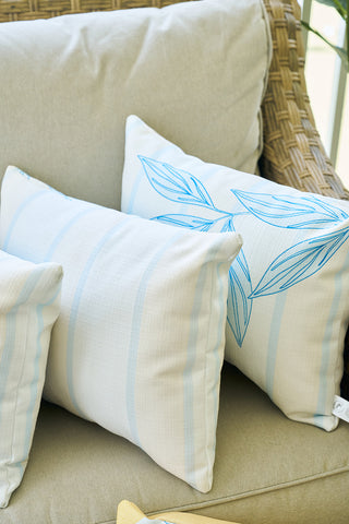 Three light-colored cushions on a wicker chair—two with pale blue stripes and one with a blue leaf pattern on cream—create a cozy setting, perfectly complemented by the Lemons in a Line Lumbar Pillow for citrus-inspired charm.