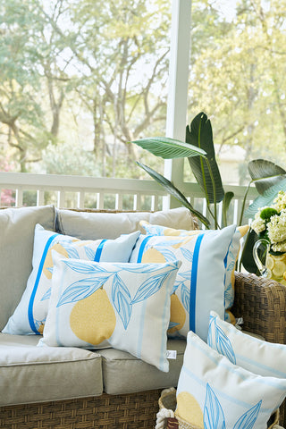 A sunlit porch with beige cushioned seating, decorated with a Lemons in a Line Lumbar Pillow featuring blue leaves. A green plant and yellow flowers are nearby, with trees visible through the window in the background.